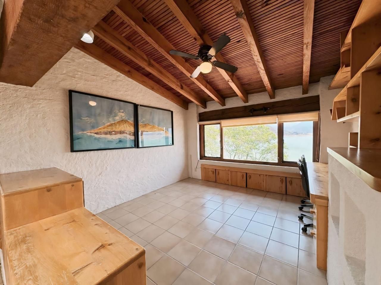 Une pièce spacieuse avec un plafond haut, des poutres apparentes, un ventilateur de plafond, une grande fenêtre offrant une belle vue et un banc intégré.