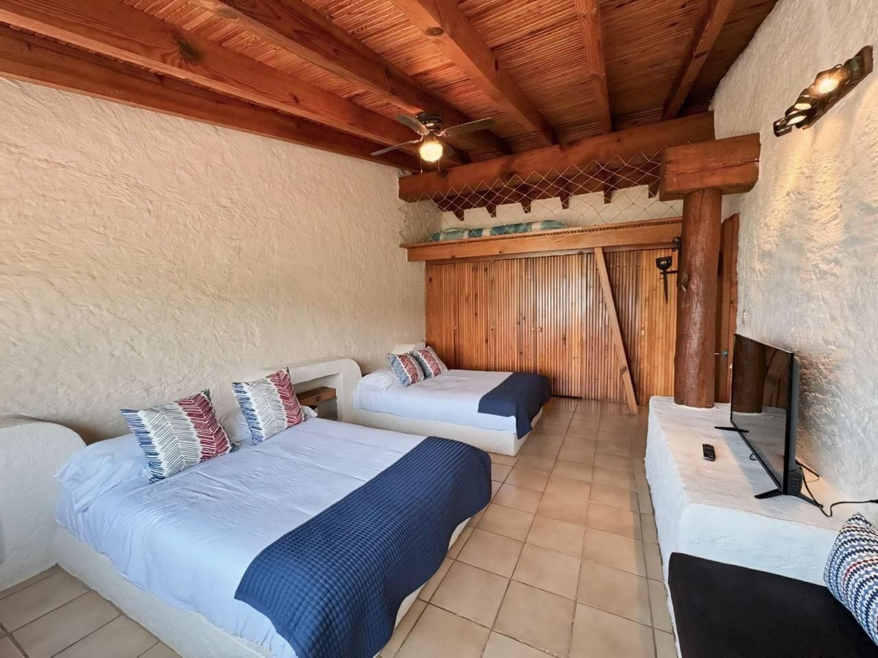 Habitación con dos camas, ventilador de techo, televisor y techo de madera.