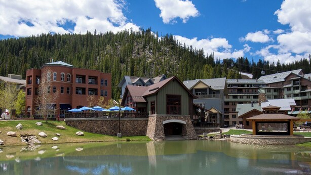 A lakeside building with a stone foundation, a wooden structure with a porch, and a backdrop of multi-story buildings and a forested mountain. - Ideal Location with 2 Queens + 2 Twins, Hot Tubs, Pool, & Clubhouse (Winter Park)