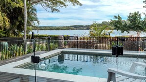 A rectangular pool with a stone statue in the center, surrounded by a wooden deck and a metal fence. - Stunning River front Cottage with pool (Dunbogan)