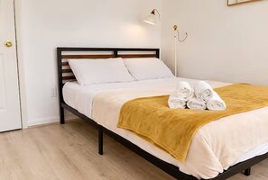 A neatly made bed with white bedding and a mustard-colored throw, a pair of rolled towels, and a modern bedside lamp. - Executive Suite Midtown (Wilmington)