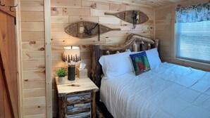 A wooden cabin room with a bed, a nightstand with a lamp, and a window with blinds. - MAPLE COTTAGE - Lakefront on Thayer Lake in Alden with WiFi, AC (Alden)