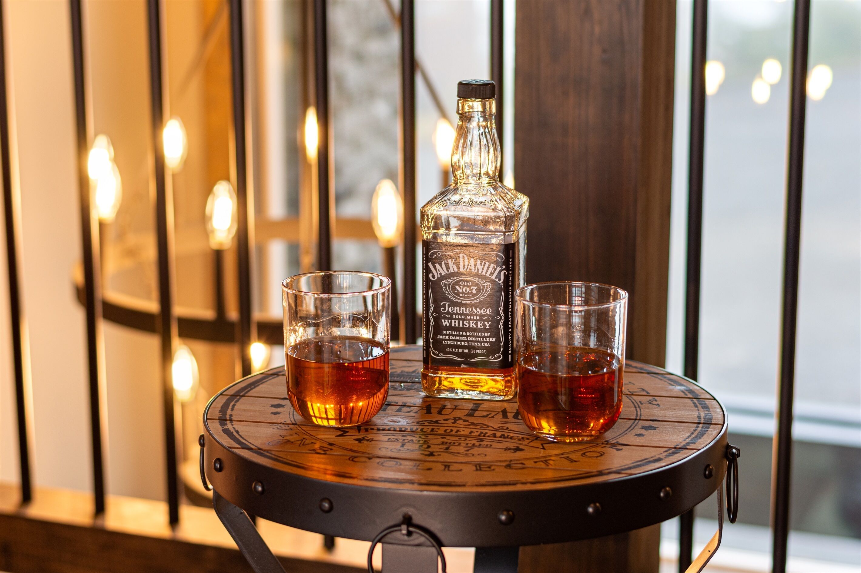 A bottle of Jack Daniel's Tennessee Sour Mash Whiskey and two glasses on a round wooden table with a metal frame.