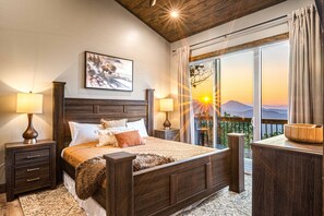 A bedroom with a wooden bed, two bedside tables with lamps, a large window with curtains, and a painting on the wall. - Wow Wow Wow a must stay cabin! (Sevierville)