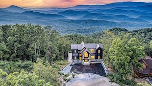 A large, multi-story house with a prominent front entrance, surrounded by dense forest and mountains in the background. - Wow Wow Wow a must stay cabin! (Sevierville)