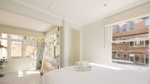 A bedroom with a large bed, a window with a view of a brick building, and a radiator. - Sugared Almond - 1 bedroom apartment Notting Hill (Greater London)