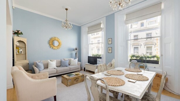 A spacious living room with a sofa, a dining table with placemats, a chandelier, and a view of buildings through large windows. - Sugared Almond - 1 bedroom apartment Notting Hill (Greater London)