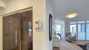 Een modern interieur met een schuifglazen deur naar een kamer, een kleine witte console tafel met een plant en een grijze fauteuil in een goed verlicht woongedeelte met grote ramen.