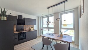 Een moderne keuken met donkere kastjes, een houten aanrechtblad en een inbouwoven. Er is een eethoek met een tafel en stoelen, en grote ramen zorgen voor natuurlijk licht.