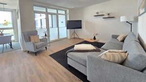 Een moderne woonkamer met een grijze bank, een grijze fauteuil, een flatscreen-tv en een eethoek met een tafel en stoelen.
