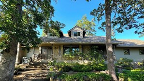 Uma casa de dois andares com uma escadaria de pedra que leva à porta da frente, cercada por vegetação exuberante e árvores.