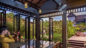 A modern bar area with a glass top, wooden chairs, and a view of a lush garden and traditional architecture. - Villa Phum Khnar (Siem Reap)