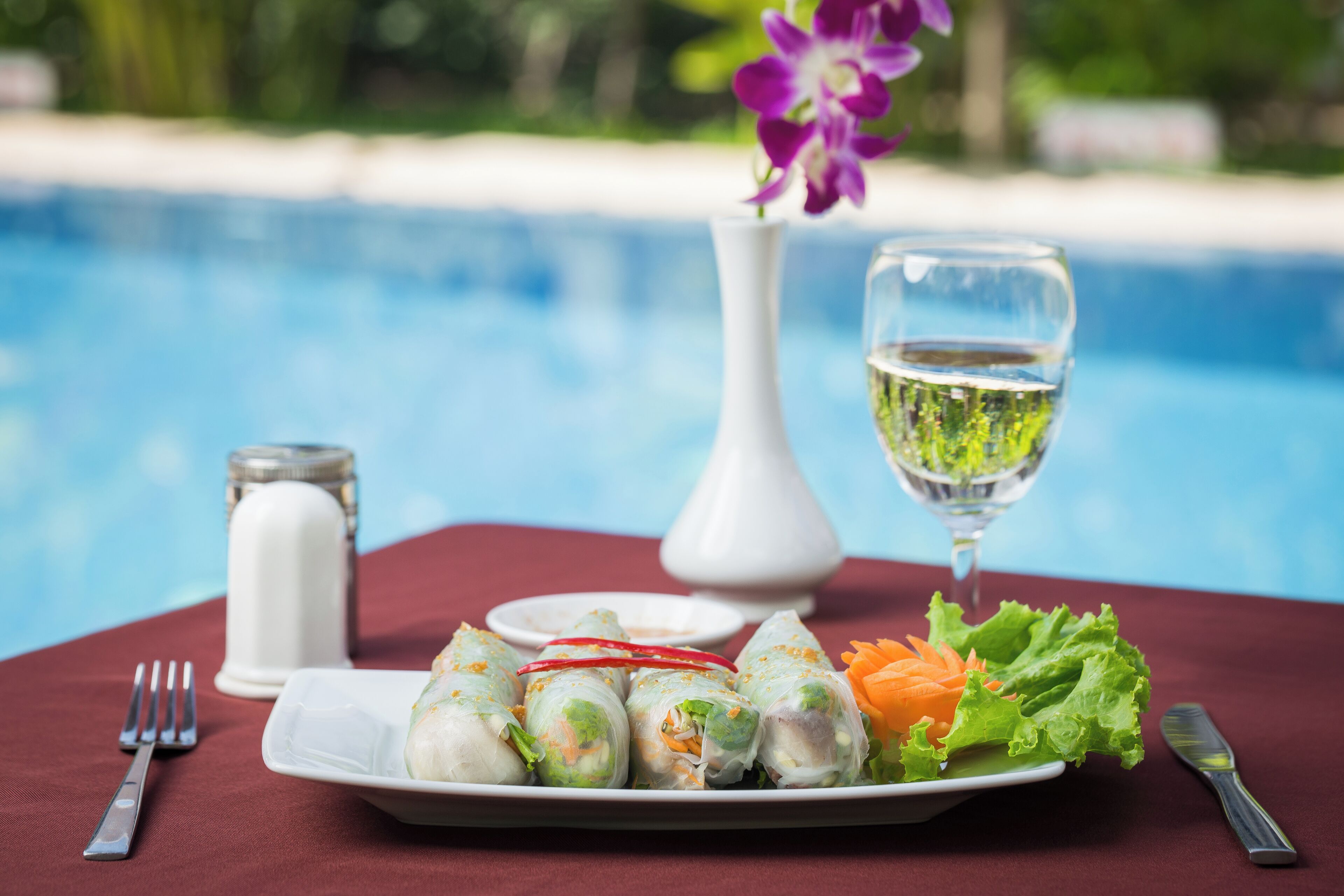 Una mesa de comedor con un plato de rollitos primavera, una copa de vino blanco y un jarrón con una flor morada.