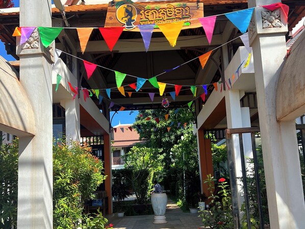 A covered walkway with colorful triangular flags, a wooden sign with text, and lush greenery. - Villa Phum Khnar (Siem Reap)