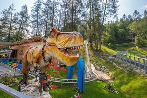 Una exposición de dinosaurios con un modelo grande de dinosaurio, un edificio de madera que dice "Alpine Coaster" y una vía de montaña rusa.