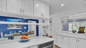 Une cuisine moderne avec des meubles blancs, un îlot central agrémenté d’un saladier de fruits, et un coin repas avec un mur bleu et des œuvres d’art.