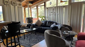 A modern living room with a black leather sofa, a glass coffee table, and a red armchair. There is a fireplace, a dining area with a table and chairs, and large windows with curtains. - 3-Bedrooms in Idyllwild - AC, 
Jacuzzi, Sauna, Fitness-Game Room,  Epic Views (Idyllwild)
