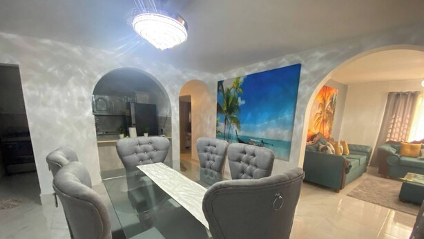 A dining area with a glass table and grey upholstered chairs, a living area with a sofa and a painting, and a kitchen in the background. - 6B Santiago Harmonic Apt Especially For You (Santiago de los Caballeros)