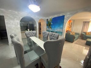 Un comedor con mesa de vidrio y sillas tapizadas de gris, una sala de estar con sofá y un cuadro, y una cocina al fondo.