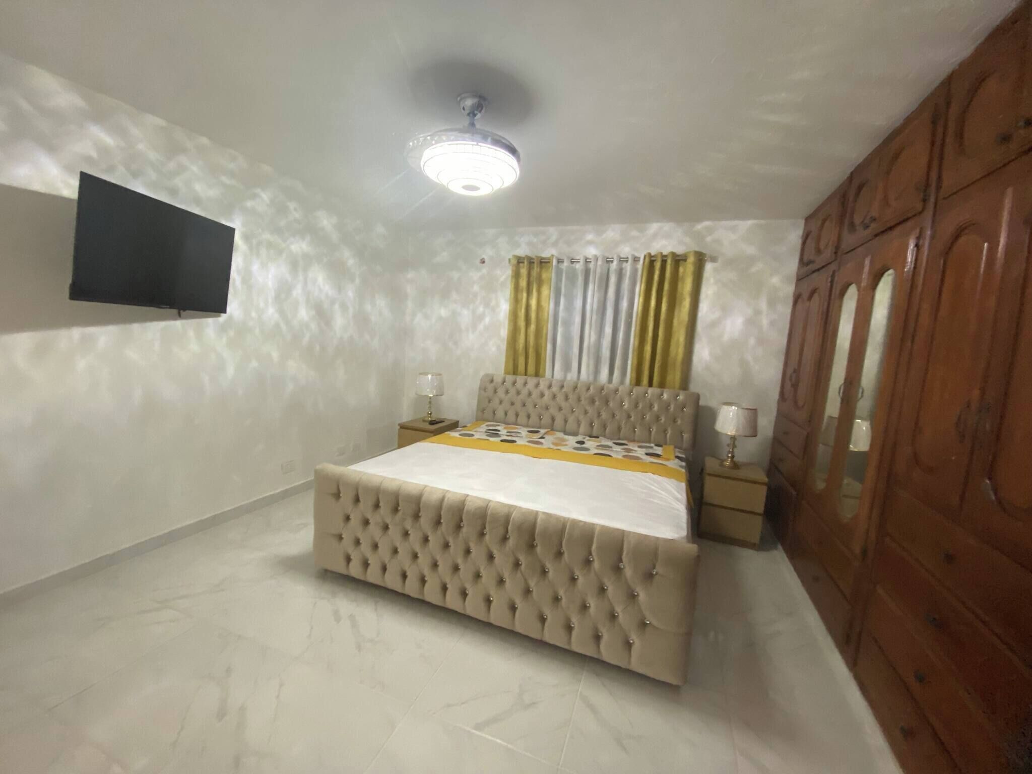 Ein Schlafzimmer mit einem Bett, einem an der Wand montierten Fernseher, einem Deckenventilator und einem Holzschrank.