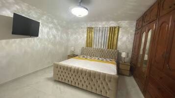 A bedroom with a bed, a TV mounted on the wall, a ceiling fan, and a wooden wardrobe.
