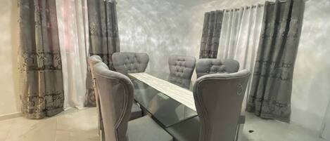 A dining room with a glass table and four upholstered chairs, a patterned rug, and curtains with a floral design.