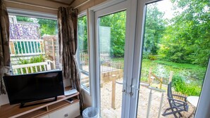 A room with a TV on a wooden stand, a sliding glass door leading to an outdoor area with a garden and a pond. - No 27 A. Field - Coghurst Hall Hastings (Hastings)
