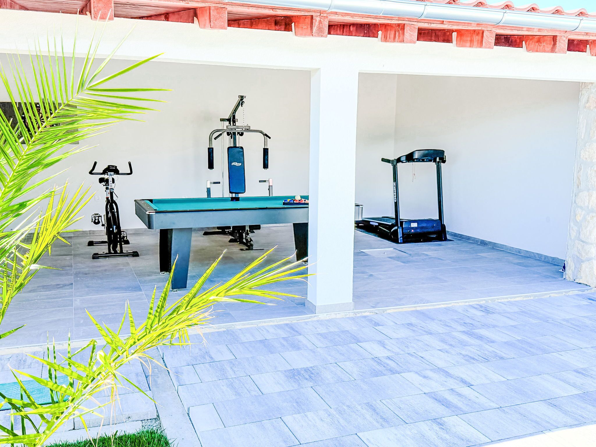 A covered outdoor area with exercise equipment including a rowing machine, a stationary bike, and a treadmill.