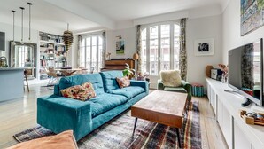 Un salon spacieux avec un canapé bleu, une table basse et un piano. On y trouve une bibliothèque remplie de livres et une grande fenêtre avec des rideaux.