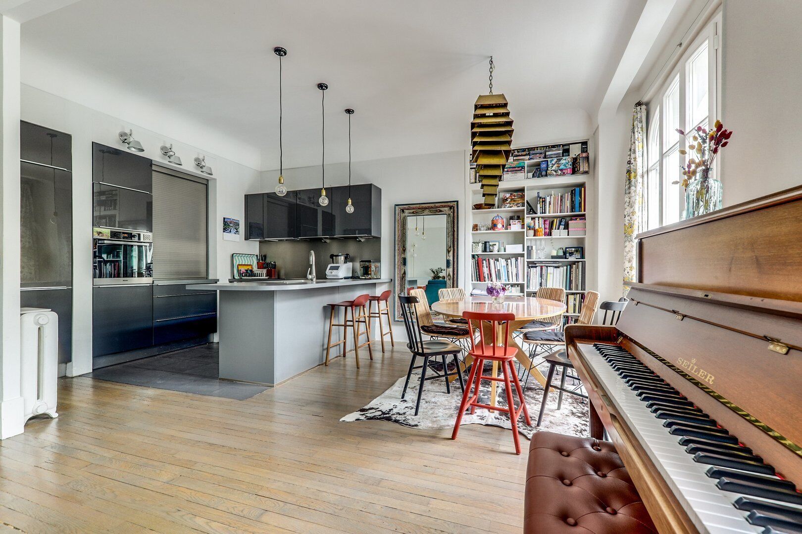 A modern kitchen with a central island, a dining area with a round table and chairs, a piano, and a bookshelf filled with books.