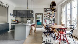 Une cuisine moderne dotée d’un îlot central, un coin repas avec une table et des chaises en bois, une bibliothèque et une grande fenêtre offrant une vue sur la ville.