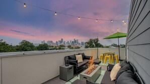 Una terraza en la azotea con una chimenea, mobiliario exterior, luces colgantes y vistas de un horizonte urbano.