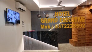 A hotel reception desk with a modern design, a mounted television displaying security footage, and a prominent sign with the hotel's name and contact number. - Hotel Grand Executive  (Mumbai)