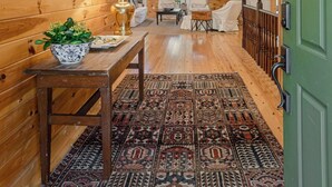 Een houten blokhutinterieur met een hoge plafond, een ster-vormige hanglamp, een eettafel met een schaal bloemen en een kamerkleed met patroon.