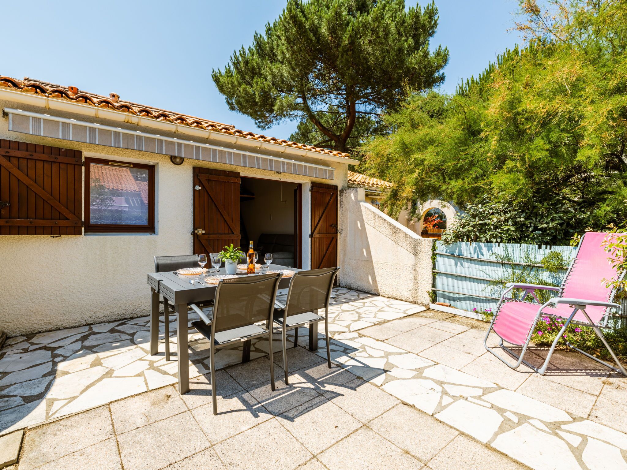 Eine Terrasse im Freien mit Tisch und Stühlen, einer Holztür und Blick auf Baum und Zaun.
