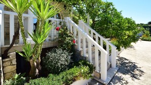 Een wit gebouw met een stenen gevel, een witte trap met balustrades, en weelderige begroeiing, waaronder een palmboom en bloeiende planten.