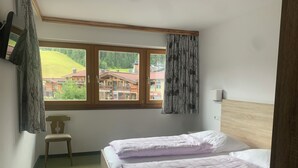 A bedroom with two beds, a chair, a window with curtains, and a view of buildings outside. - Appartements Robert Bechlwirt (Kirchberg in Tirol)