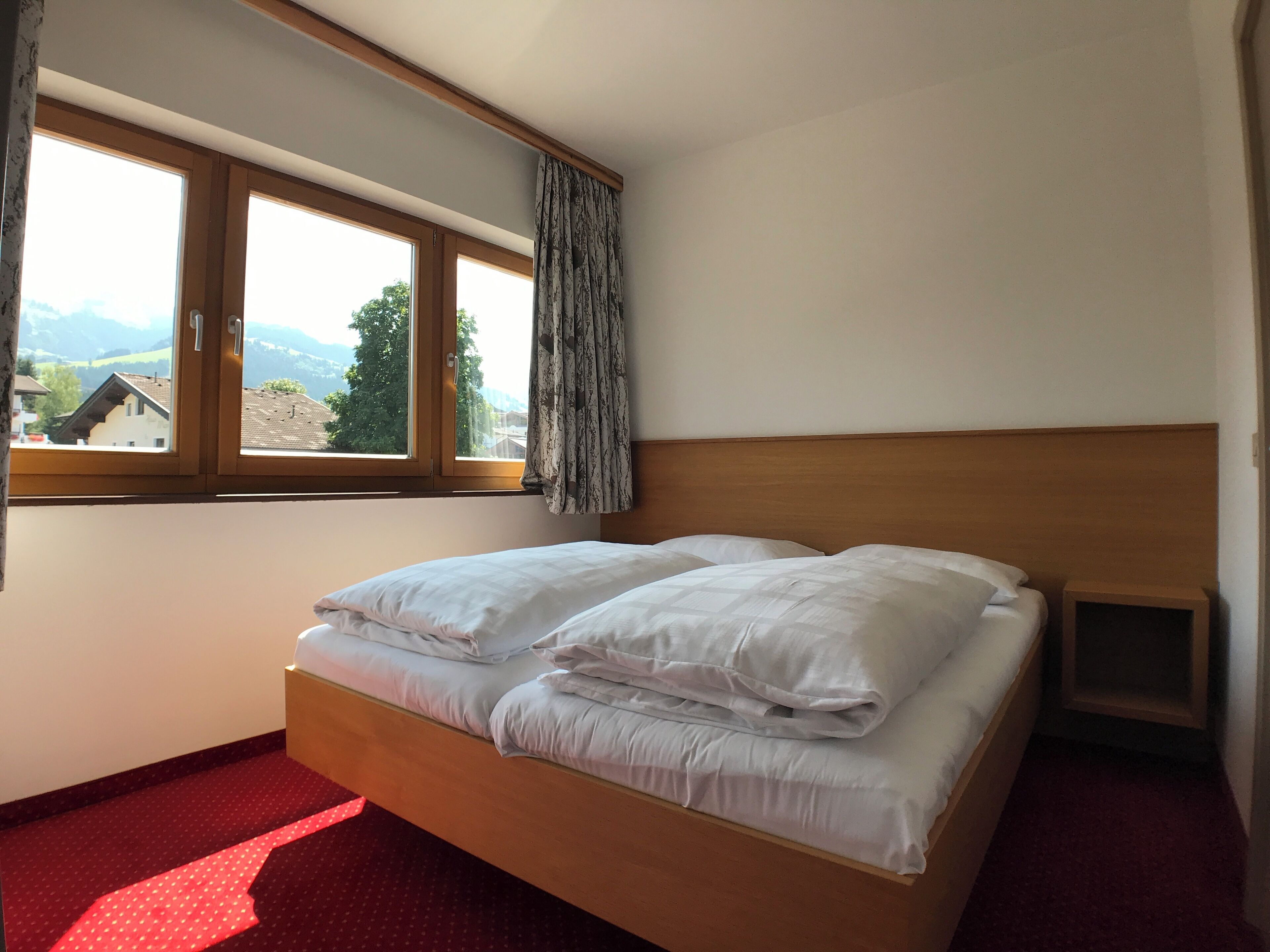 A bedroom with a wooden bed, a window with curtains, and a view of buildings and mountains.