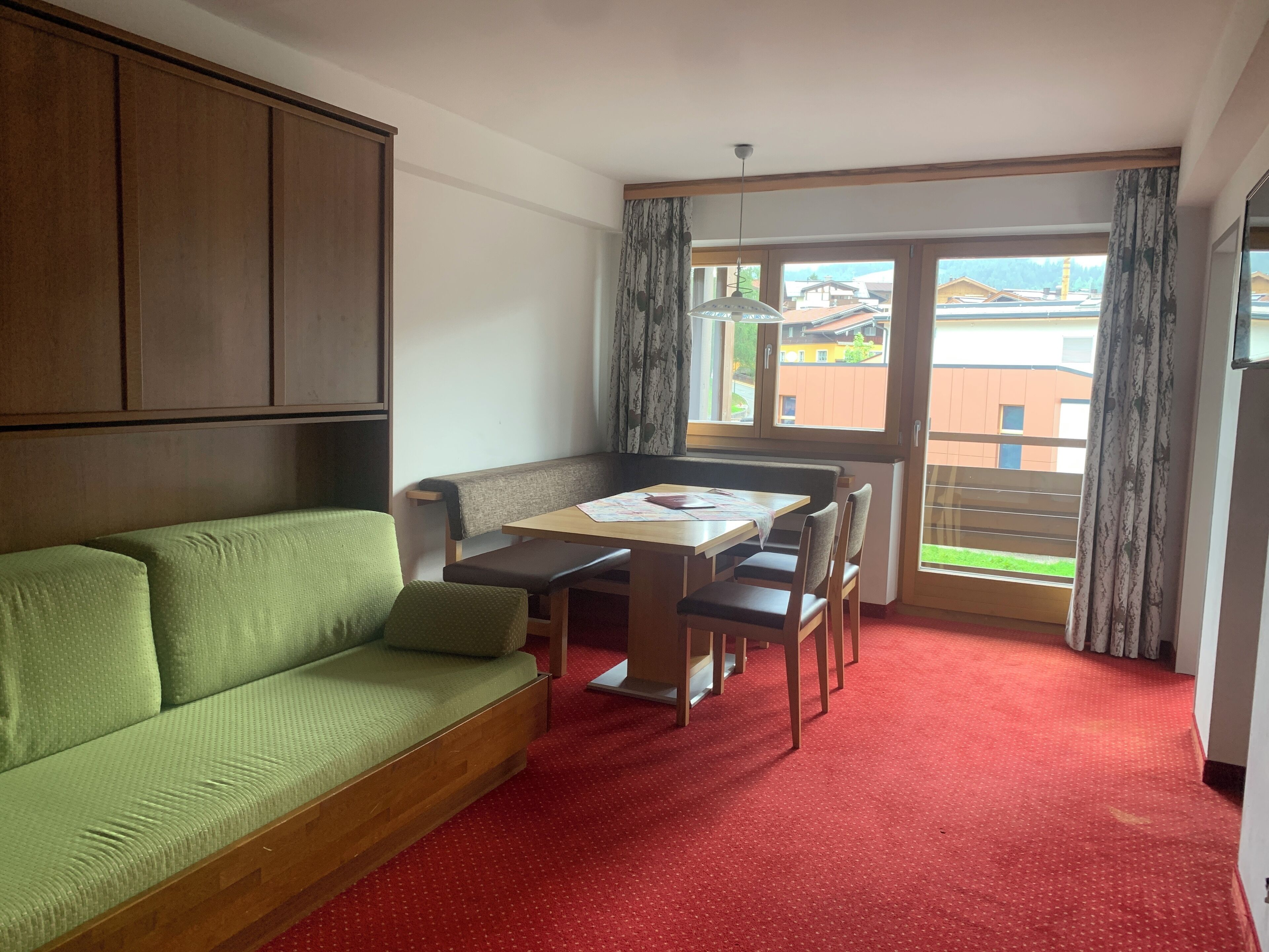 A room with a green sofa, a wooden table with chairs, a red carpet, and a view of buildings through a window with curtains.
