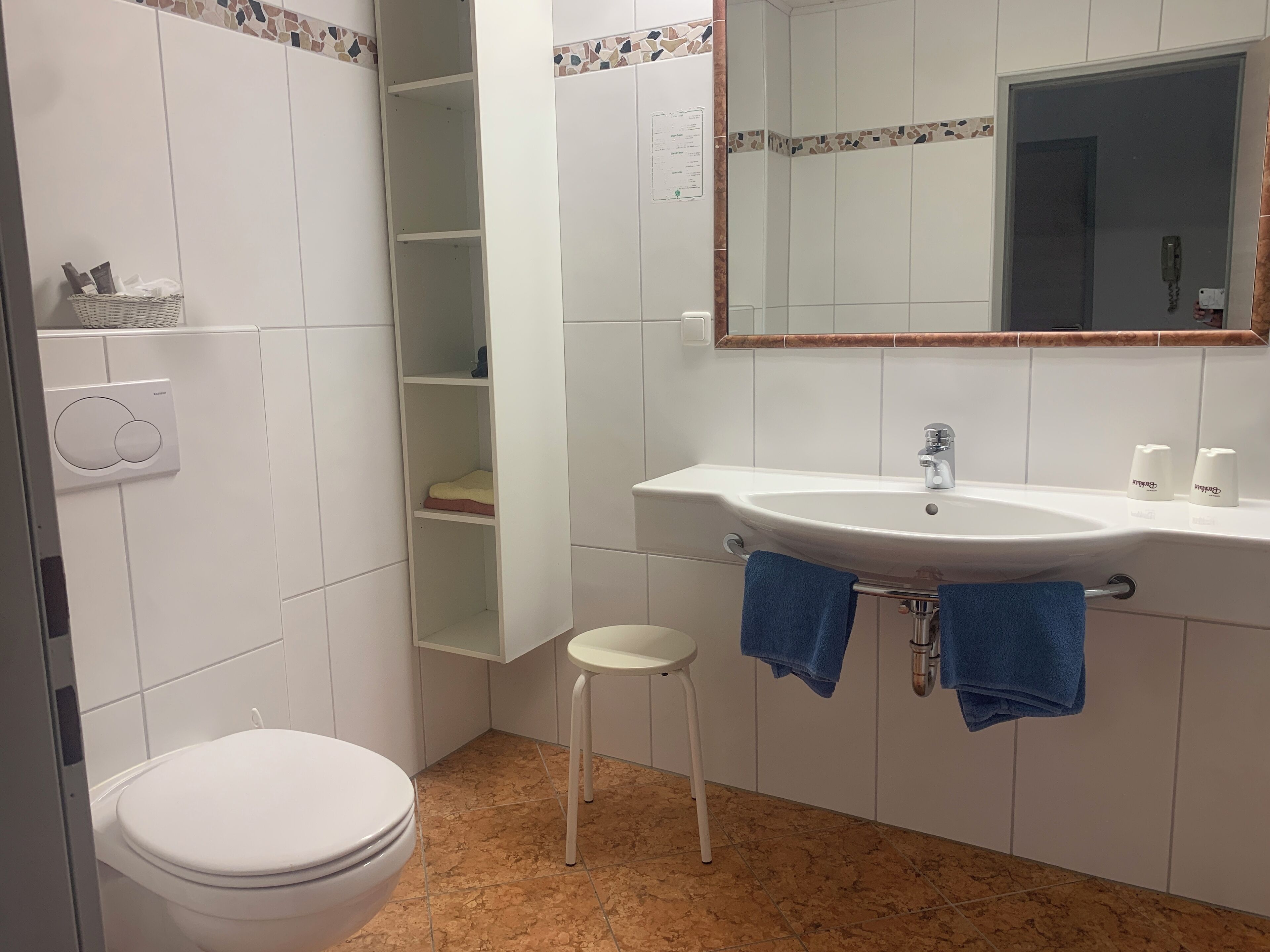 A bathroom with a white toilet, a white sink with two towels, a small stool, and a tiled floor.