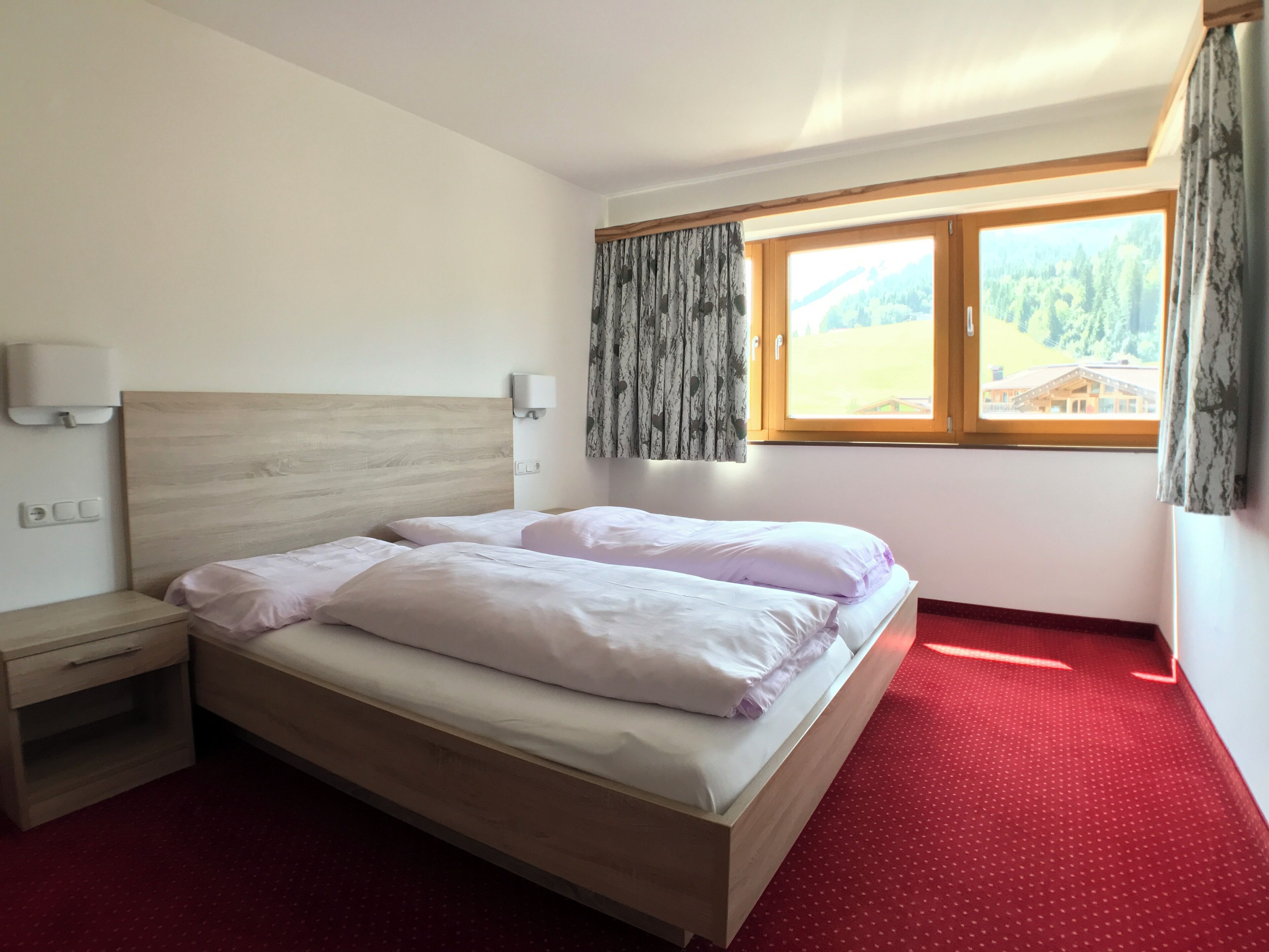 A hotel room with a wooden bed, a nightstand, a red carpet, a window with a view of a forest and a building, and a wall-mounted light fixture.