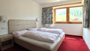 A hotel room with a wooden bed, a nightstand, a red carpet, a window with a view of a forest and a building, and a wall-mounted light fixture.