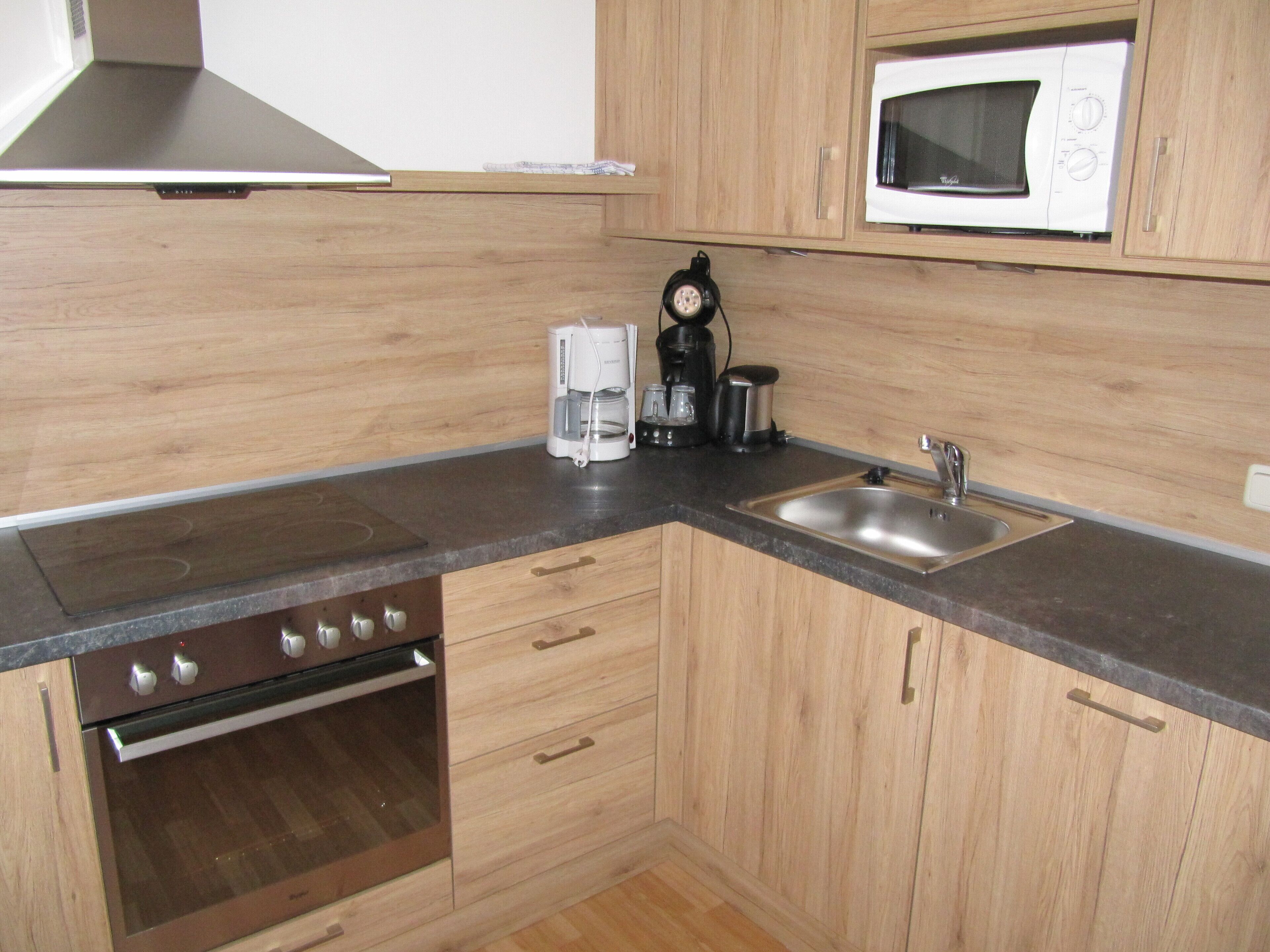 Una cocina compacta con armarios de madera, un lavamanos de acero inoxidable, un microondas, una cafetera y un horno.