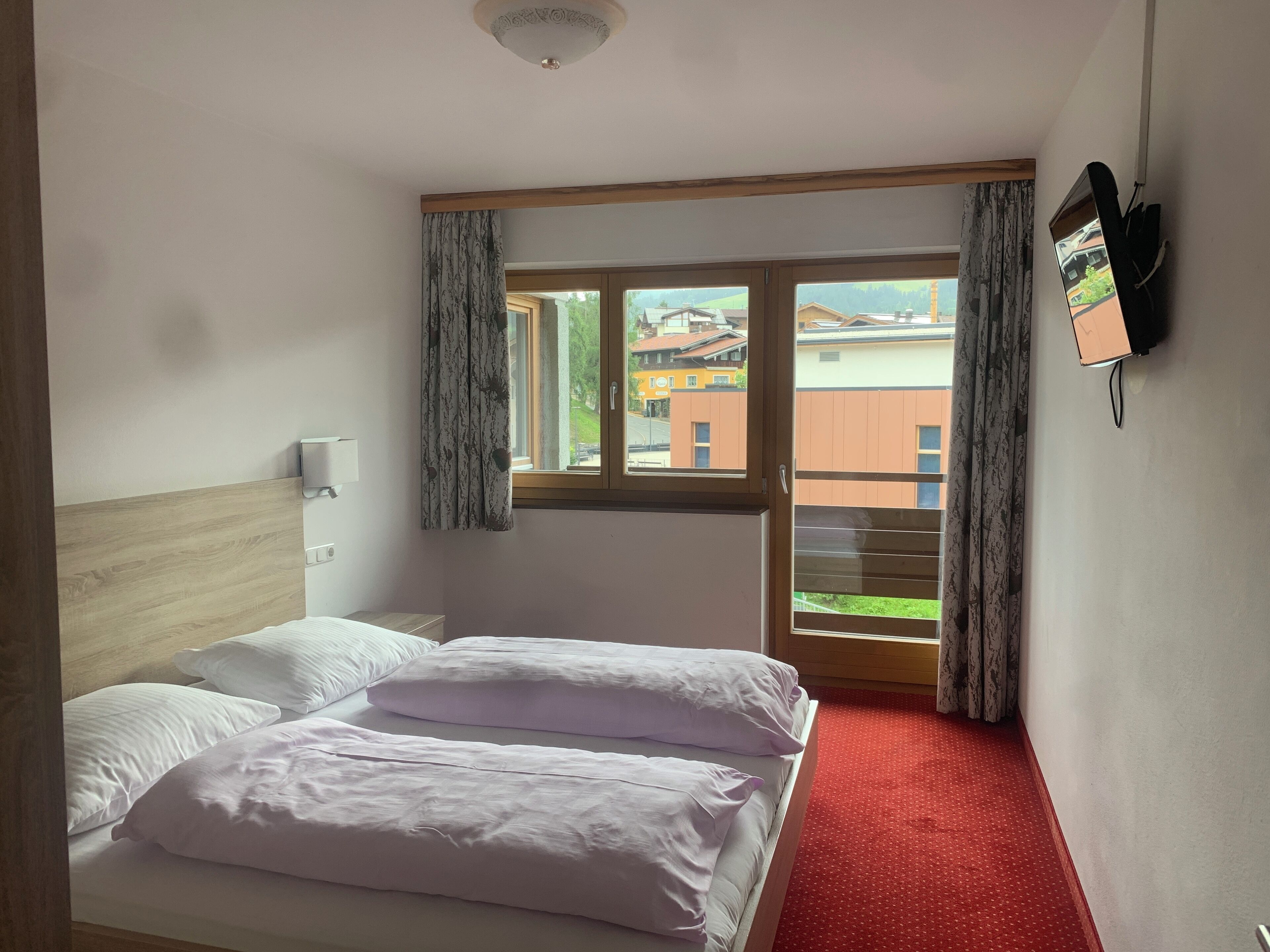 Une chambre d’hôtel avec un lit, une fenêtre avec des rideaux, un téléviseur fixé au mur et une moquette rouge.