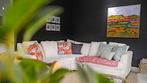 Una sala de estar con un sofá blanco, cojines estampados, una mesa de centro de vidrio y un cuadro en la pared.