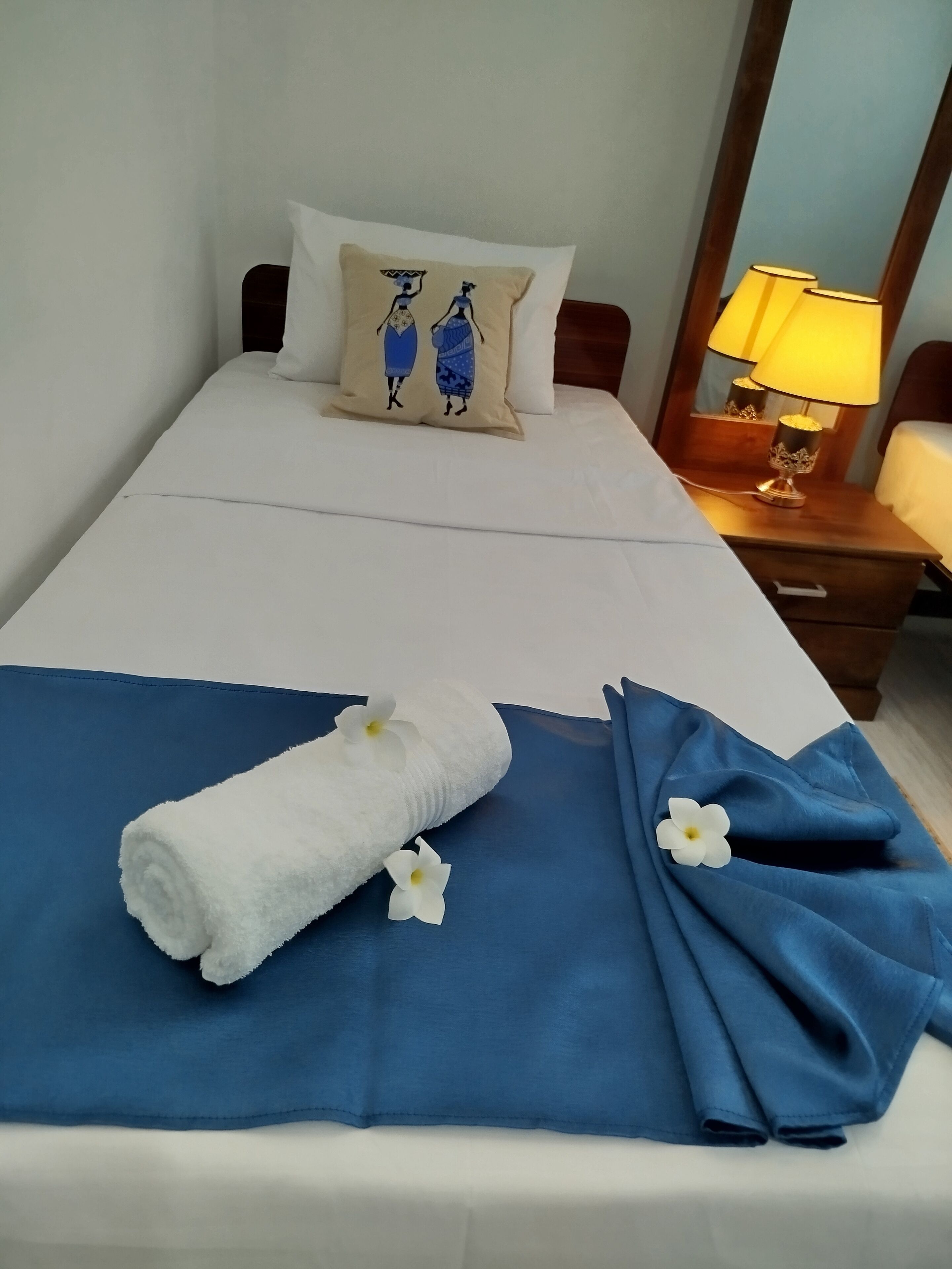 Una cama bien tendida con un edredón azul, una toalla blanca con flores y una almohada con un diseño cultural.