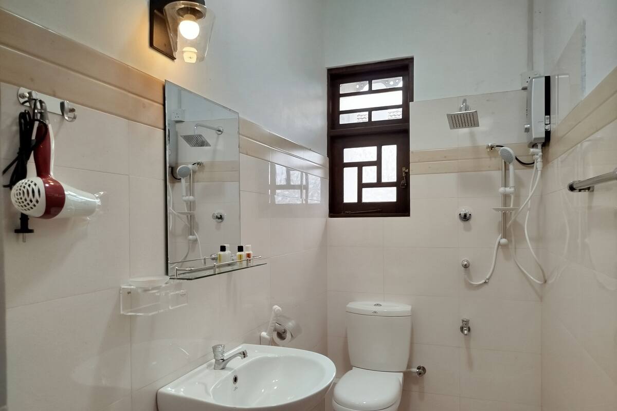 Egy fürdőszoba, amelyben fehér mosdókagyló, WC és zuhanyzó található. A mosdókagyló felett van egy tükör, a zuhanyzó felett pedig egy kis ablak.
