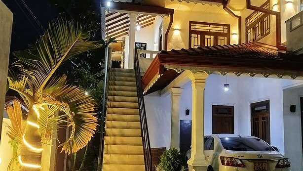 A two-story house with a staircase leading to the upper floor, a parked car, and potted plants. - Cosy White Palace  (Negombo)