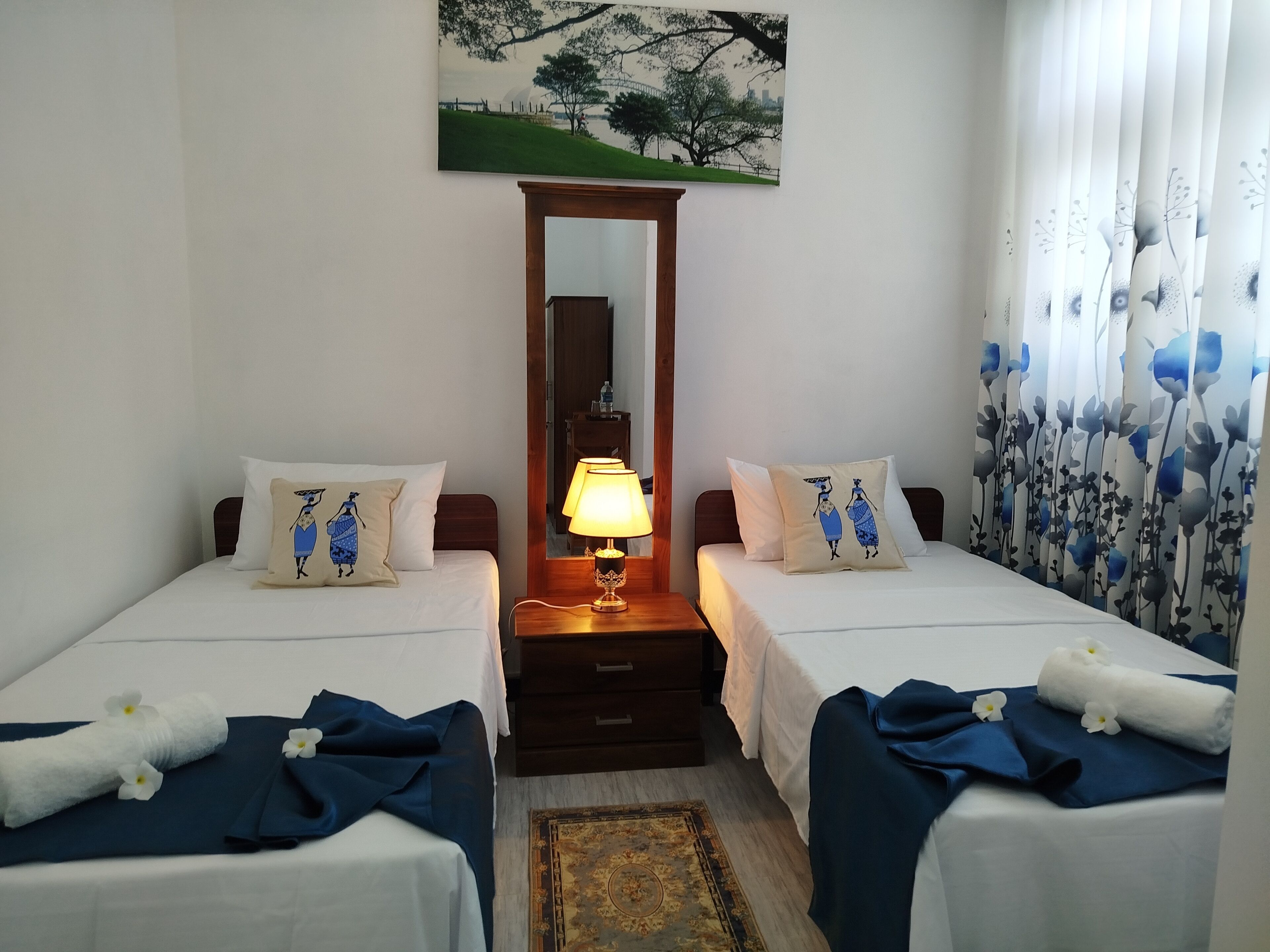 A hotel room with two single beds, a nightstand with a lamp, a mirror, and a painting on the wall.