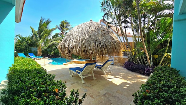 A pool area with lounge chairs, a thatched umbrella, and tropical plants. - Wonderful 1-bedroom Apartment 3 km From Eagle Beach (Noord)